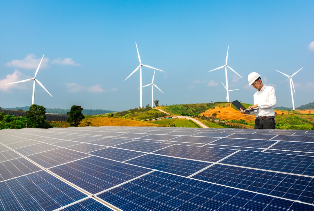 Ein Techniker mit Helm und Laptop steht auf einer Solaranlage. Hinter ihm sind Windräder zu sehen.