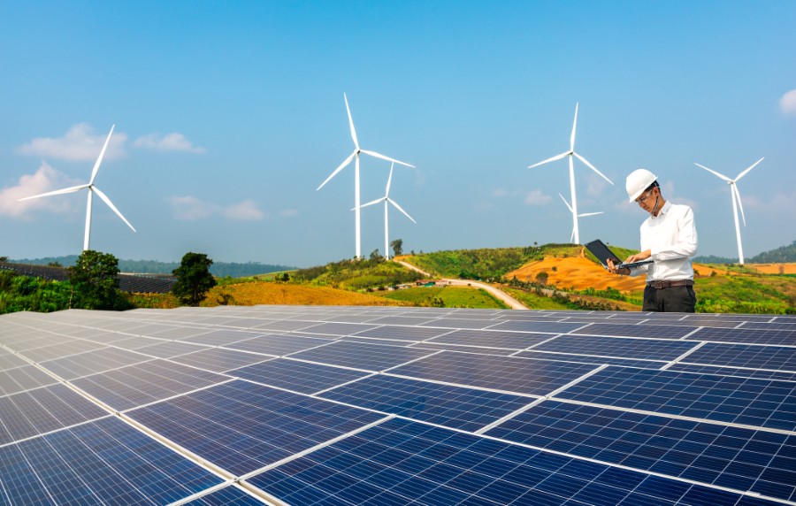 Ein Techniker mit Helm und Laptop steht auf einer Solaranlage. Hinter ihm sind Windräder zu sehen.